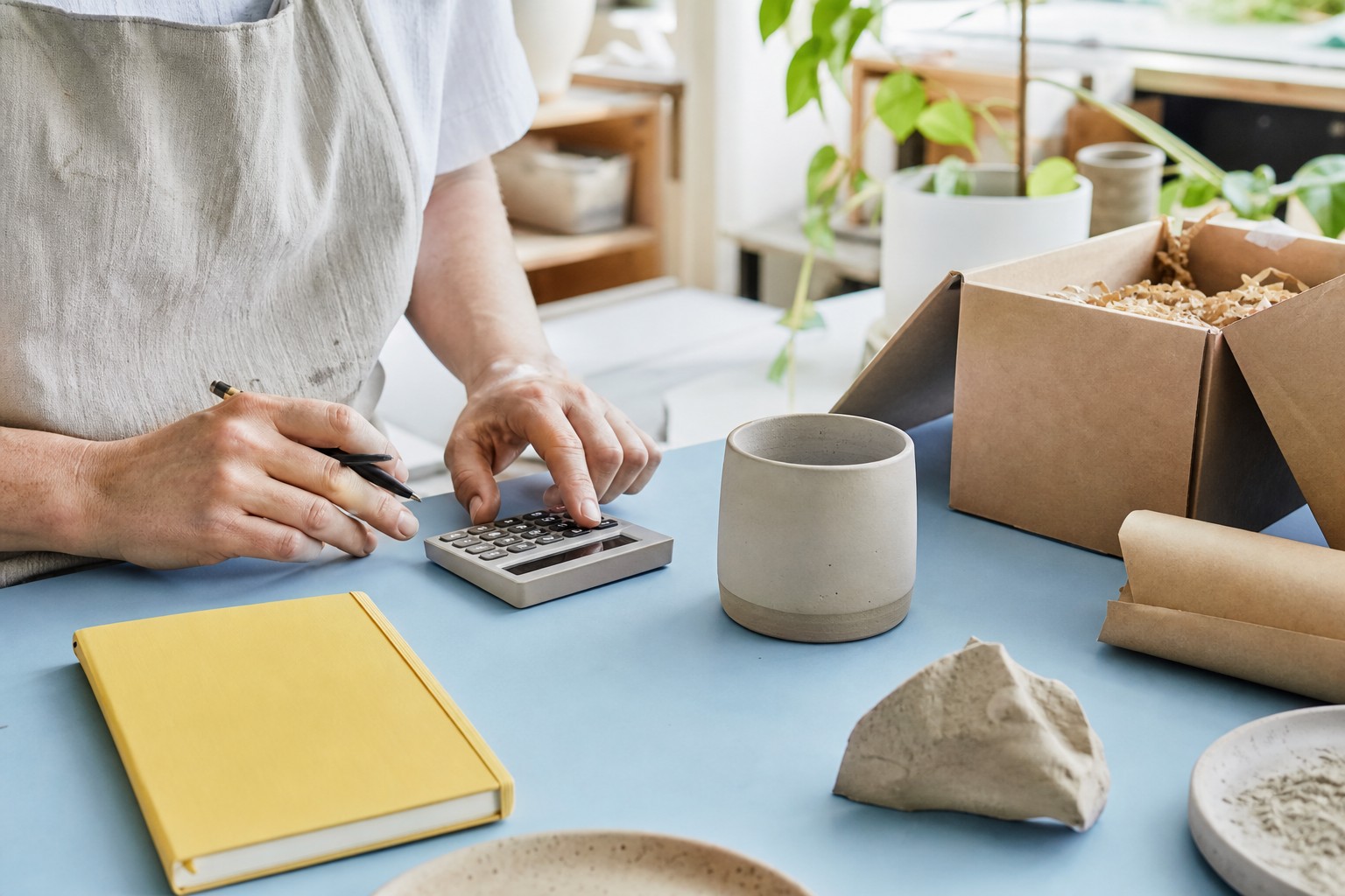 Person berechnet am Studiotisch den Preis einer Keramiktasse mit Taschenrechner und Materialproben